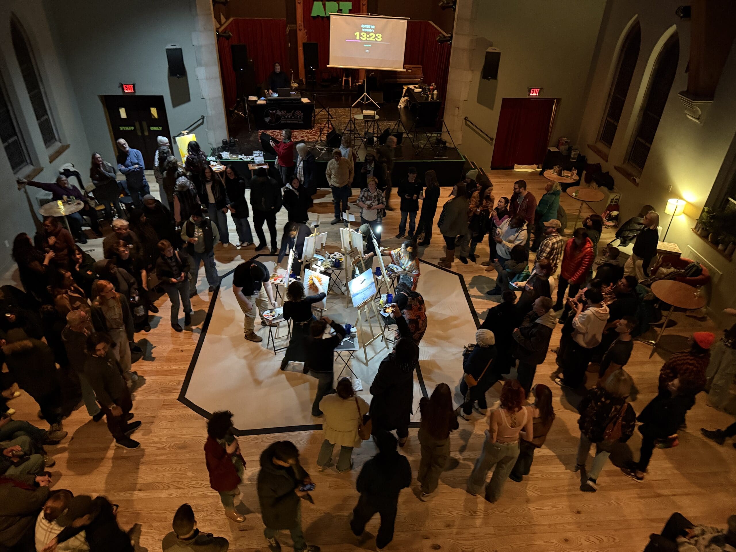 Image of a room full of people circled around 6 artists painting at easels