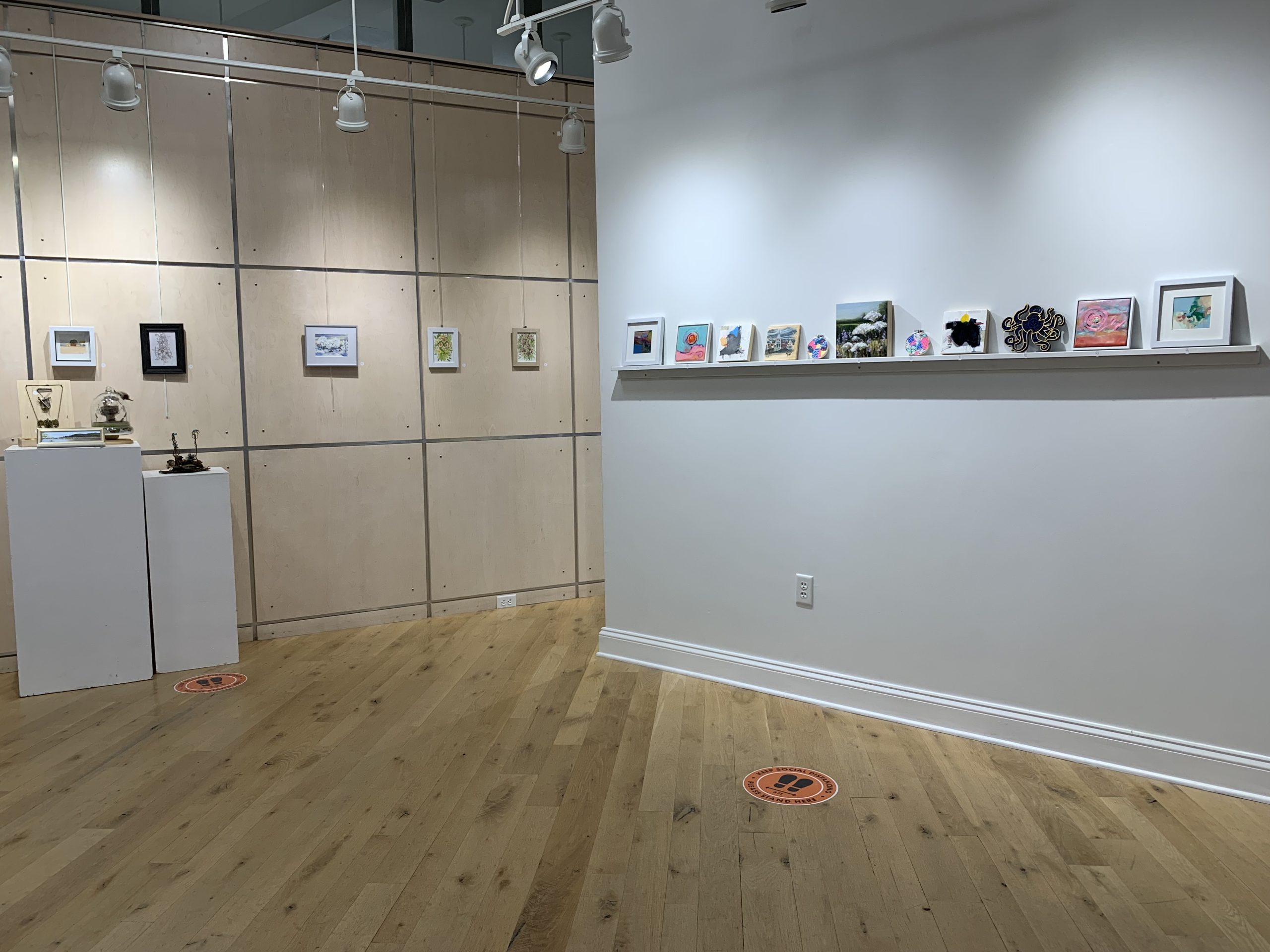 gallery showing 100 under 100 exhibit. artworks in a row sitting on a shelf with pedestals to the left with art