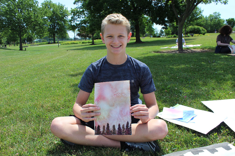 teen boy smiling and holding up a watercolor landscape while sitting in the grass